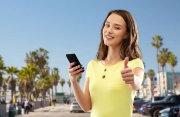 technology and people concept - smiling young woman or teenage girl in yellow t-shirt with smartphone showing thumbs up over venice beach background in california. teenage girl with smartphone showing thumbs up