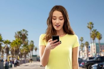 technology and people concept - smiling young woman or teenage girl in blank yellow t-shirt using smartphone over venice beach background in california. teenage girl using smartphone over venice beach