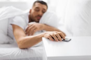 technology and people concept - close up of sleepy young man reaching for smartphone on bedside table from bed at home in morning. sleepy young man reaching for smartphone in bed