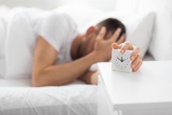 morning and people concept - close up of sleepy young man in bed reaching for alarm clock on bedside table at home. close up of man in bed reaching for alarm clock