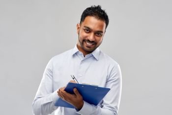 business, office worker and people concept - smiling indian businessman writing to clipboard over grey background. smiling indian businessman writing to clipboard