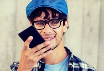leisure, technology, communication and people concept - smiling hipster man using voice command recorder or calling on smartphone at street wall. man using voice command or calling on smartphone