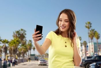 technology and people concept - smiling young woman or teenage girl in blank yellow t-shirt taking selfie by smartphone over venice beach background in california. teen girl takes selfie by cell over venice beach