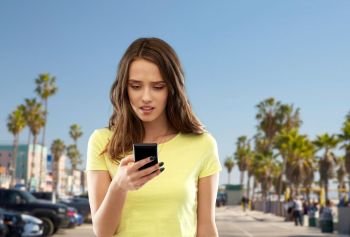 technology, cyberbullying and people concept - confused young woman or teenage girl in blank yellow t-shirt using smartphone over venice beach background in california. teenage girl using smartphone over venice beach