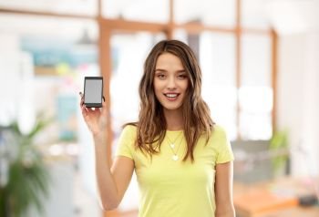 technology and people concept - smiling young woman or teenage girl in yellow t-shirt holding smartphone with blank screen over office background. young woman or teenage girl holding smartphone