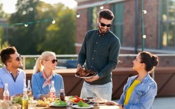 leisure and people concept - happy party host offering meat to his friends at bbq on rooftop in summer. friends at bbq party on rooftop in summer
