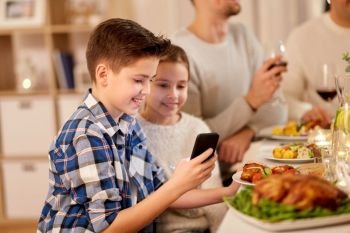 technology, holidays and people concept - happy boy with sister using smartphone having family dinner party at home. boy with sister using smartphone at family dinner