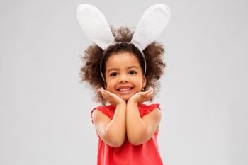 childhood, party props and easter concept - happy little african american girl wearing bunny ears headband posing over grey background. happy little girl wearing easter bunny ears posing