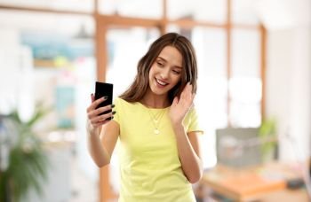 technology and people concept - smiling young woman or teenage girl in blank yellow t-shirt having video call smartphone and waving hand over office background. smiling teenage girl having video call smartphone