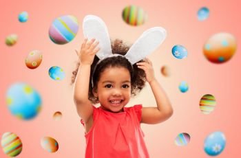 childhood, party props and easter concept - happy little african american girl wearing bunny ears headband over colored eggs on living coral background. happy little girl wearing easter bunny ears
