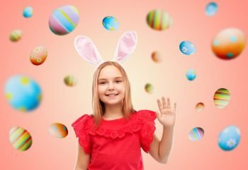easter, holidays and childhood concept - happy girl wearing bunny ears headband waving hand over colored eggs on living coral background. happy girl wearing easter bunny ears waving hand