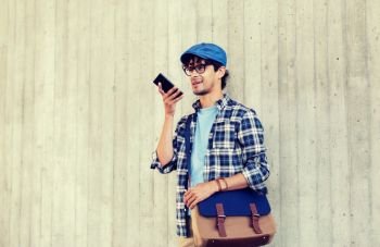 leisure, technology, communication and people concept - smiling hipster man with shoulder bag using voice command recorder or calling on smartphone at street wall. man using voice command or calling on smartphone