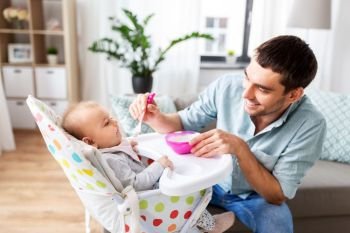 family, food, eating and people concept - happy father feeding little baby daughter sitting in highchair with puree by spoon at home. father feeding happy baby in highchair at home