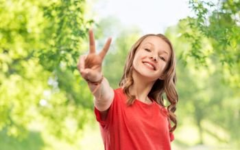 gesture and people concept - smiling teenage girl with long wavy hair in red t-shirt showing peace hand sign over green natural background. smiling teenage girl in red t-shirt showing peace