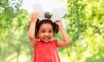 childhood, party props and easter concept - happy little african american girl wearing bunny ears headband over green natural background. happy little girl wearing easter bunny ears