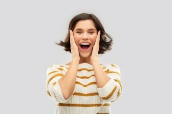 emotion, expression and people concept - happy impressed young woman in striped pullover holding to her face over grey background. impressed woman in pullover holding to her face