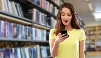technology and people concept - smiling young woman or teenage girl in blank yellow t-shirt using smartphone over library background. young woman or teenage girl using smartphone