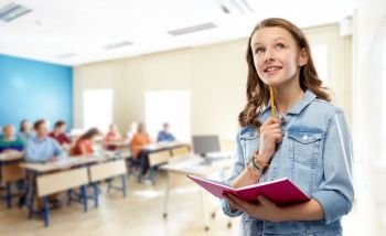 education, school and people concept - teenage student girl in denim jacket with diary or notebook and pencil thinking over classroom background. teenage student girl with notebook at school