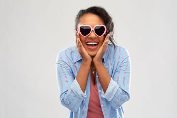 summer, valentine’s day and emotion concept - happy african american young woman in heart-shaped sunglasses over pink background. african american woman in heart-shaped sunglasses