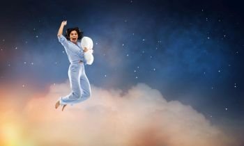 fun, people and bedtime concept - happy young woman full of energy in blue pajama holding pillow and jumping over starry night sky background. happy woman in blue pajama jumping with pillow