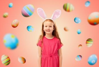 easter, holidays and childhood concept - happy red haired girl wearing bunny ears headband over living coral background and colored eggs. happy red haired girl wearing easter bunny ears