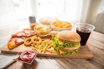 fast food and unhealthy eating concept - close up of double hamburger or cheeseburger, deep-fried squid rings, french fries, pizza and cola drink on wooden board. close up of fast food and cola drink on table