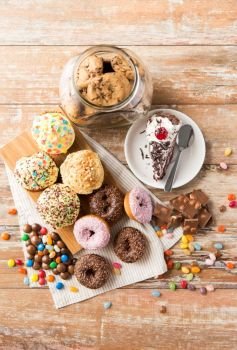 sweets and food concept - close up of glazed donuts, chocolate, drop candies, oatmeal cookies and piece of cake on wooden table. close up of different sweets on table