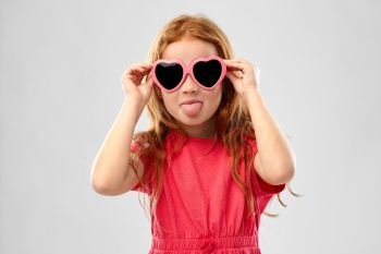 childhood, valentines day and summer concept - naughty red haired girl with heart shaped sunglasses showing tongue over grey background. naughty red haired girl in heart shaped sunglasses