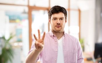 count and people concept - smiling young man showing three fingers over office room background. young man showing three fingers over office