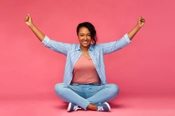 success, winning gesture and people concept - happy young african american woman celebrating victory and sitting on floor over pink background. happy african american woman celebrating success