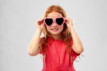 childhood, valentine’s day and summer concept - smiling red haired preteen girl in heart shaped sunglasses over grey background. smiling red haired girl in heart shaped sunglasses