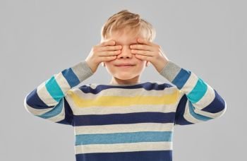 childhood, vision and seeing concept - little boy in striped pullover closing his eyes by hands over grey background. boy in striped pullover closing his eyes by hands