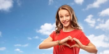 valentine’s day, love and charity concept - smiling teenage girl with long hair in red t-shirt making hand heart gesture over blue sky and clouds background. smiling teenage girl in red making hand heart