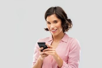 technology and people concept - happy smiling young woman in striped shirt using smartphone over grey background. young woman in striped shirt using smartphone