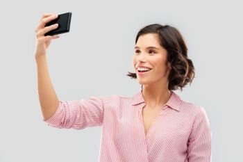 fashion, style and people concept - happy smiling young woman in striped shirt taking selfie by smartphone over grey background. smiling young woman taking selfie by smartphone