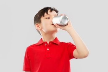 childhood, expressions and people concept - portrait of smiling little boy in red polo t-shirt drinking soda from tin can over grey background. boy in red t-shirt drinking soda from tin can