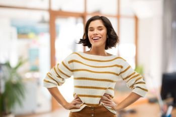 people concept - happy smiling young woman in striped pullover with hands on hips over office room background. smiling woman in pullover with hands on hips