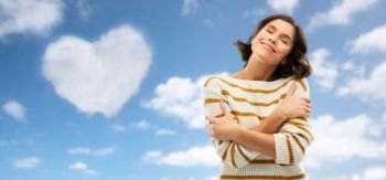 valentine’s day, love people concept - happy pleased young woman in striped pullover with closed eyes hugging herself over blue sky and cloud in shape of heart background. woman hugging herself over heart shaped cloud