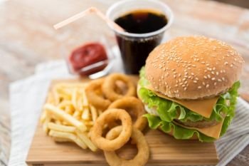 fast food and unhealthy eating concept - close up of double hamburger or cheeseburger, deep-fried squid rings, french fries, cola drink and ketchup on wooden board. close up of hamburger and other fast food