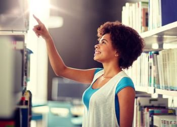 education, high school, university, learning and people concept - happy smiling african american student girl pointing finger to book on shelf at library. african student girl pointing to book at library