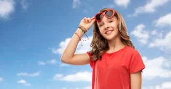 valentine’s day, love and people concept - smiling pretty teenage girl wearing bangles on hand in red heart shaped sunglasses over blue sky and clouds background. happy teenage girl in red heart shaped sunglasses