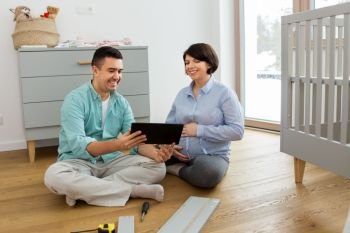 pregnancy, family and nursery concept - happy middle-aged man and his pregnant wife with tablet computer assembling baby bed at home. family couple with tablet pc assembling baby bed