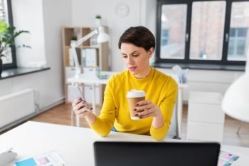 business, technology and user interface design concept - businesswoman or ui designer using smartphone and drinking takeaway coffee from paper cup at office. woman with coffee using smartphone at office