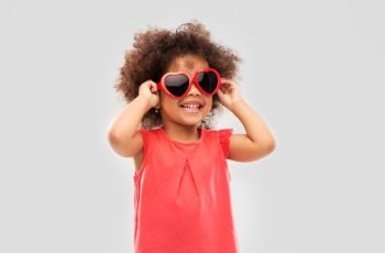 childhood, valentine’s day and summer concept - happy little african american girl in heart shaped sunglasses over grey background. african ameican girl in heart shaped sunglasses