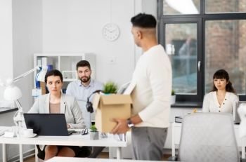 business, firing and job loss concept - sad office workers looking at fired male colleague with box of personal stuff. sad office workers looking at fired colleague