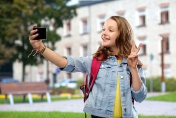 education, school and people concept - happy smiling teenage student girl with bag taking selfie by smartphone and showing peace over campus background. teenage student girl taking selfie by smartphone