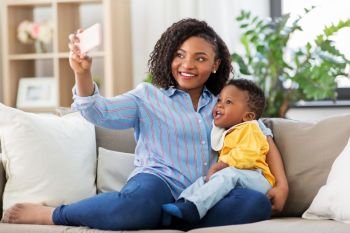 family, technology and motherhood concept - happy smiling young african american mother with little baby son taking selfie by smartphone at home. african mother with baby son taking selfie at home