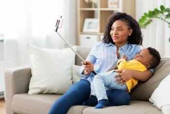 family, technology and motherhood concept - happy smiling young african american mother with little baby son taking picture by smartphone on selfie stick at home. african mother with baby son taking selfie at home