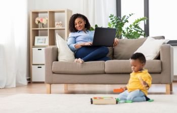 family, motherhood and people concept - happy african american mother with laptop computer looking at her little baby son playing with toy blocks kit at home. mother with laptop looking at baby with toy blocks