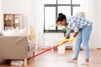 housework and housekeeping concept - african american woman or housewife with mop cleaning floor under sofa at home. african woman or housewife cleaning floor at home